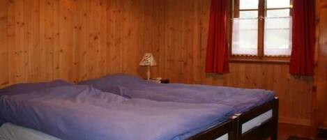 Una habitación con paredes de madera, una cama con colcha azul, una mesita de noche con lámpara y una ventana con cortinas rojas.