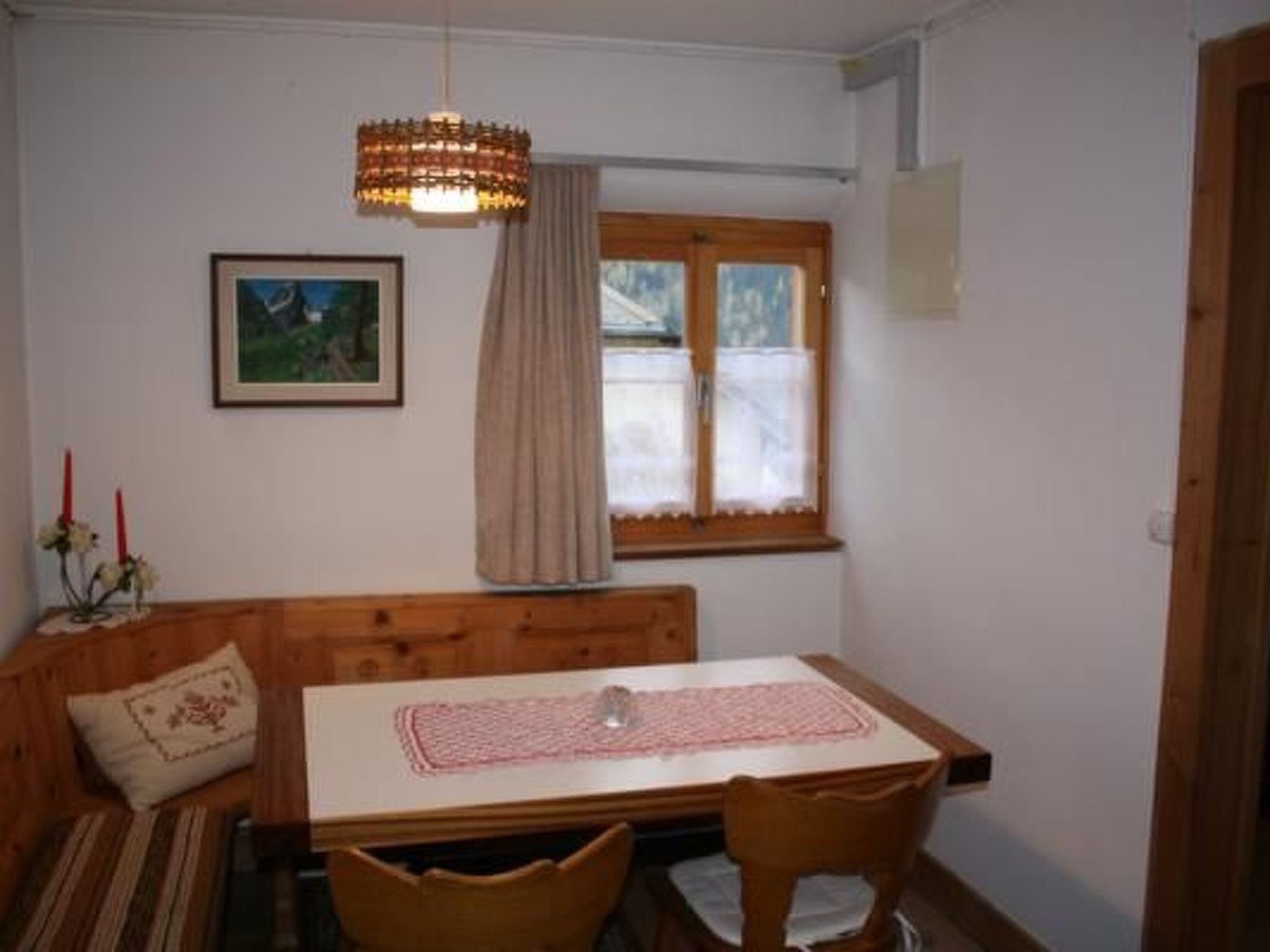 Un comedor con mesa de madera, sillas, banco, lámpara colgante, ventana con cortinas e imagen enmarcada en la pared.