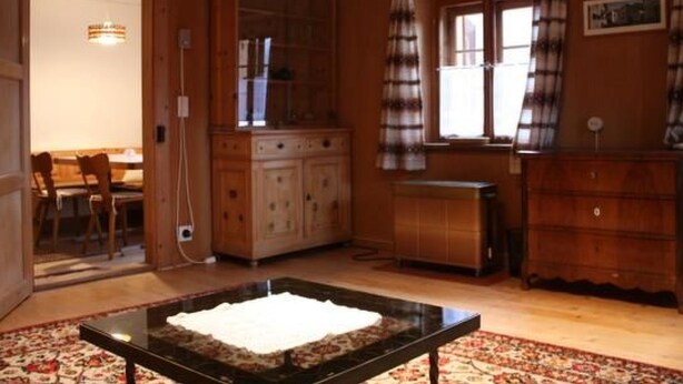 Habitació amb parets de fusta i una finestra gran amb cortines. Hi ha una taula de menjador amb cadires, un armari i una taula petita amb un objecte blanc a sobre.