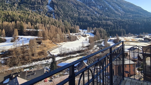 Un paysage de montagne avec un sommet enneigé, une vallée parsemée d’arbres et un bâtiment doté d’une rambarde métallique au premier plan.