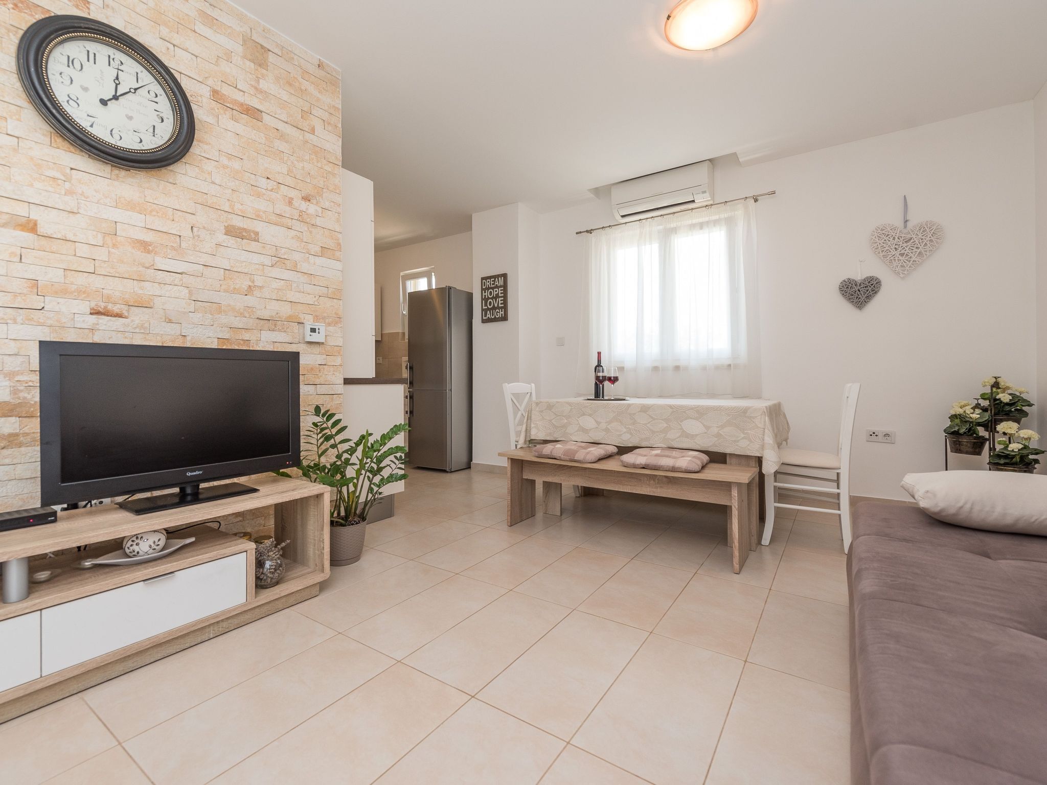 Una sala moderna con televisor de pantalla plana, pared con diseño de ladrillo, comedor con banco y cocina al fondo.