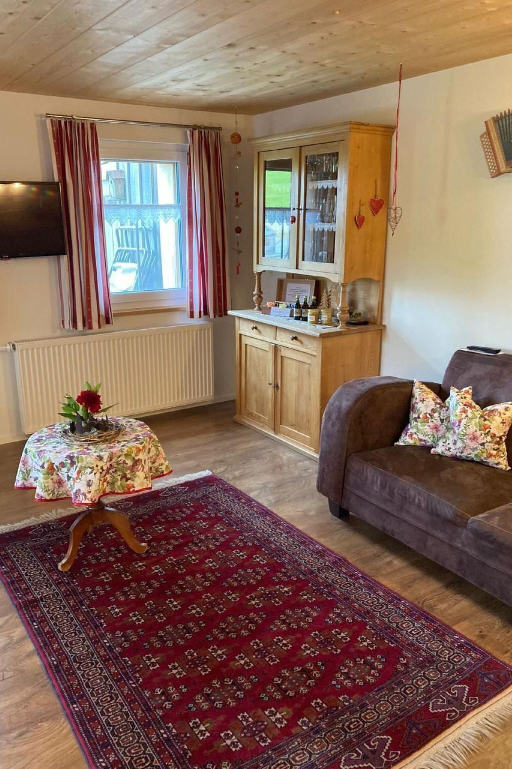 Ein gemütliches Wohnzimmer mit Holzbalkendecke, einer Couch mit floralen Kissen, einem kleinen Tisch mit blumigem Tischläufer und einem roten Teppich mit Muster.