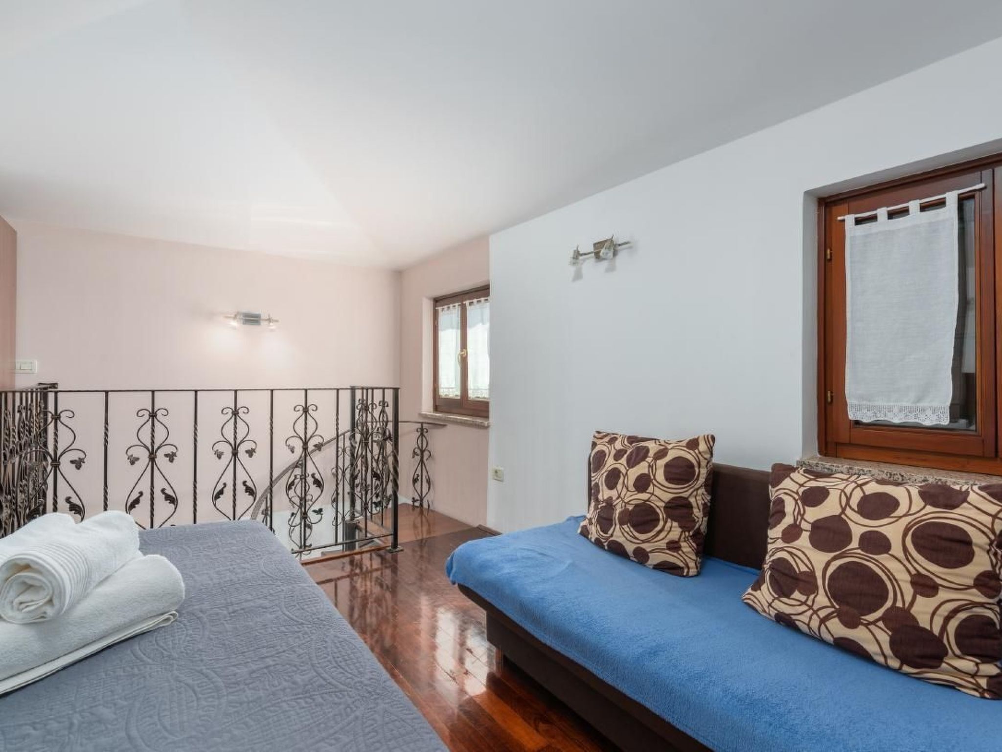 Ein Schlafzimmer mit einem Bett, einem blauen Sofa, gemusterten Kissen, einem Fenster mit Vorhang und einem Balkon mit Metallgeländer.