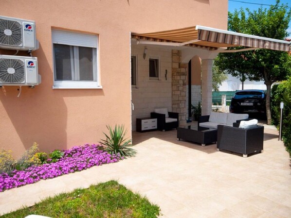 Un espace extérieur aménagé avec des canapés et une pergola, des unités de climatisation fixées au mur, et une voiture garée à proximité.
