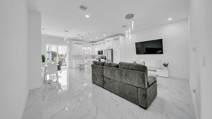 Un salón moderno y espacioso con un sofá gris, un televisor de pantalla plana en la pared y una cocina elegante al fondo.