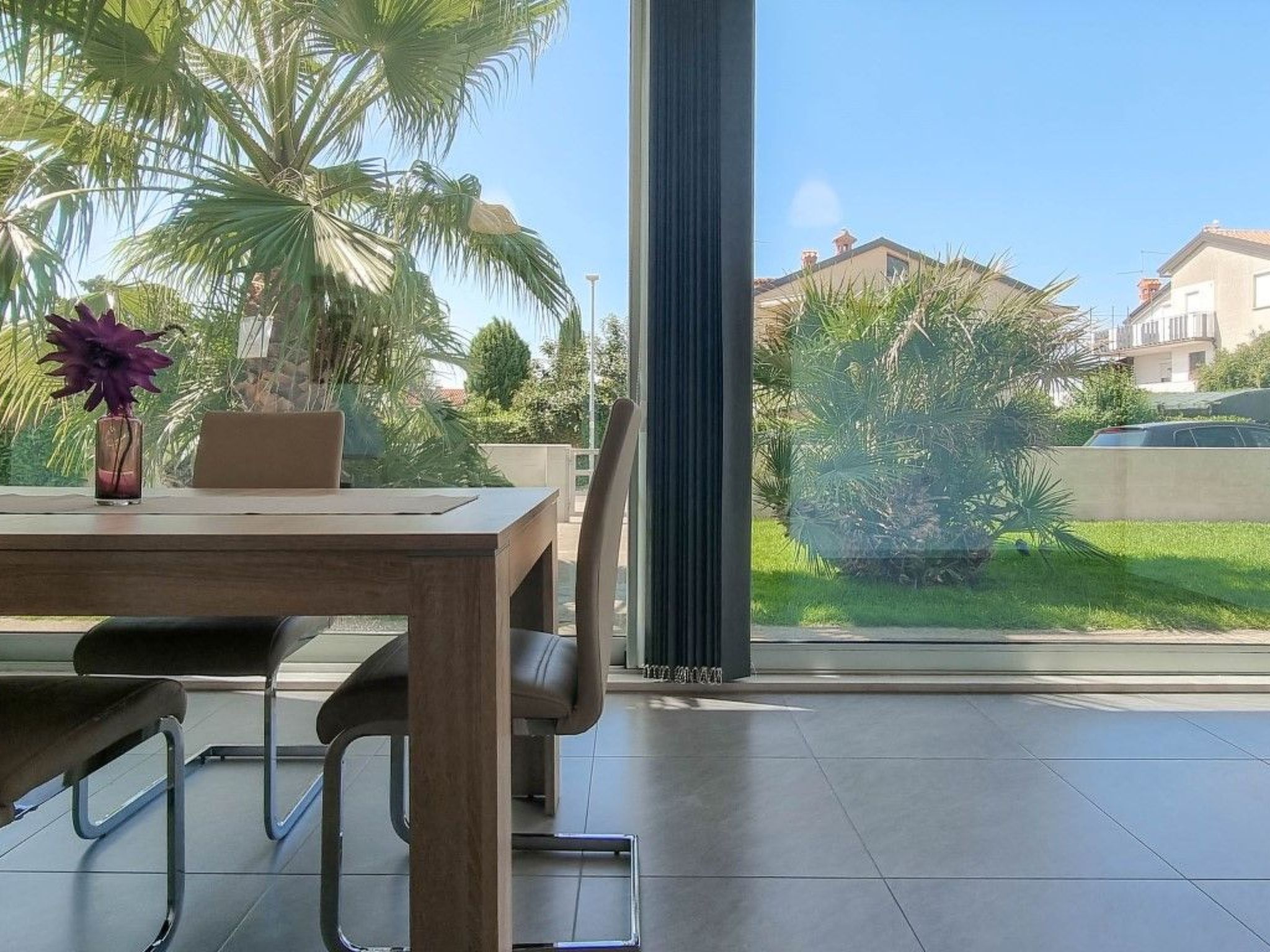 A modern dining area with a wooden table and metal chairs, a vase with purple flowers, and a view of a green garden and houses through large windows.