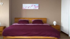 Um quarto com cama de madeira, roupa de cama roxa, decoração em formato de coração na parede e uma pequena mesa de cabeceira com abajur.