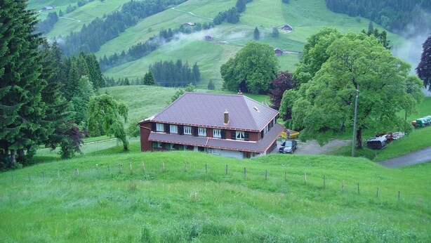 A house with a sloped roof, surrounded by lush green fields and trees, with a clear view of the surrounding hills.