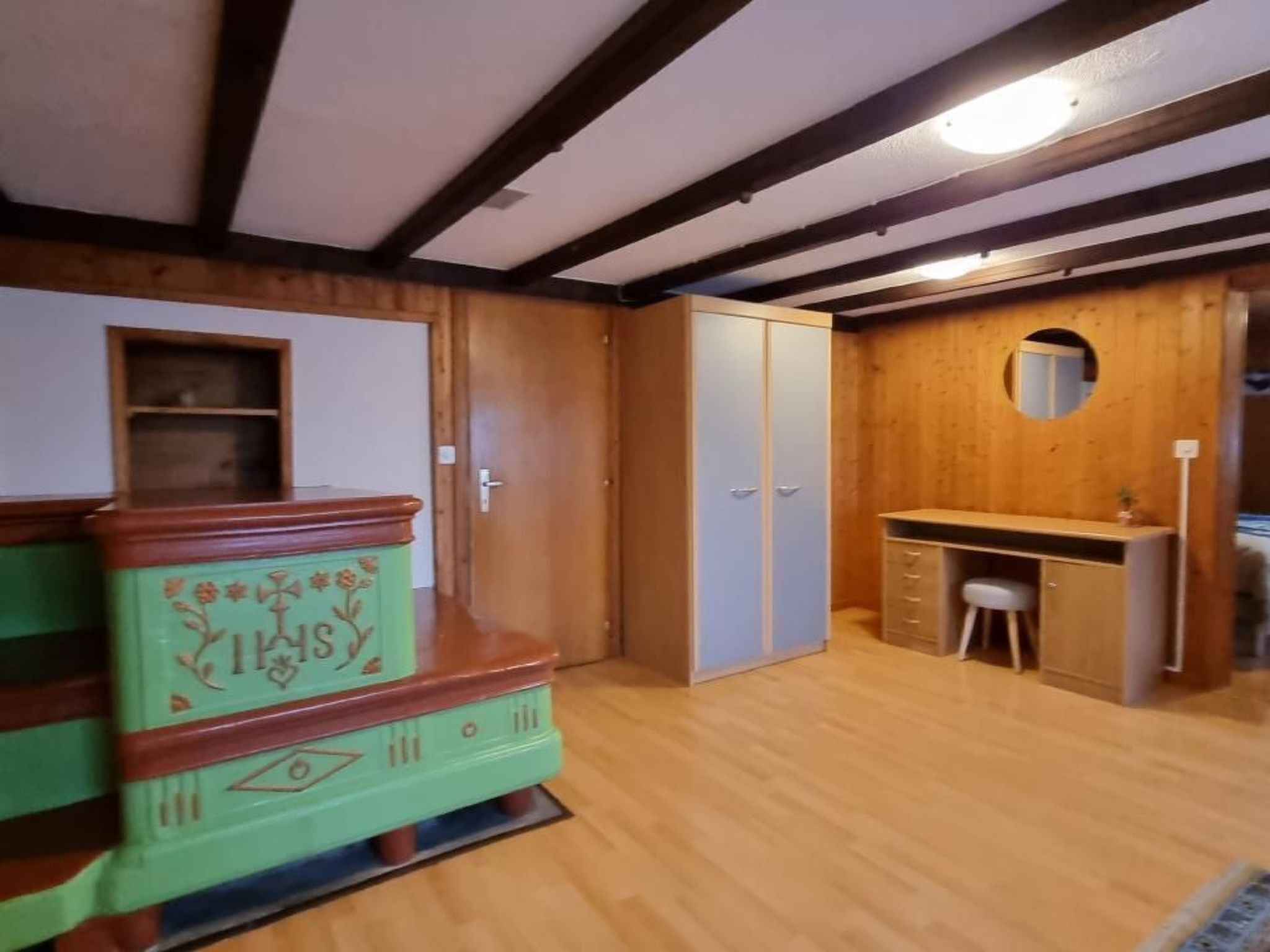 Una habitación con techo de madera, un aparador verde, un armario, un escritorio con silla y un espejo redondo pequeño.