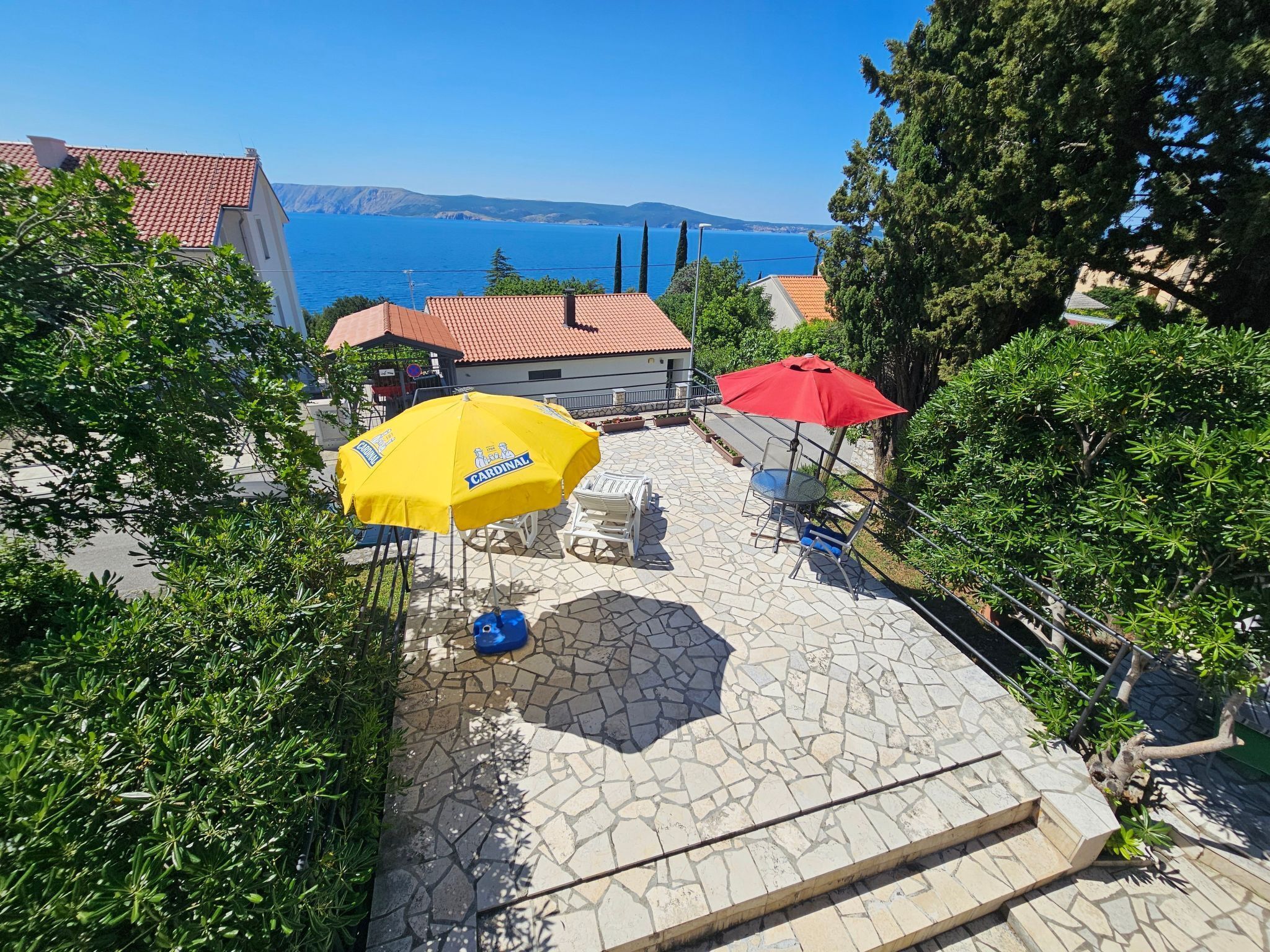 Un patio con mobiliario exterior y sombrillas, suelo de piedra y vistas al mar y a las montañas al fondo.