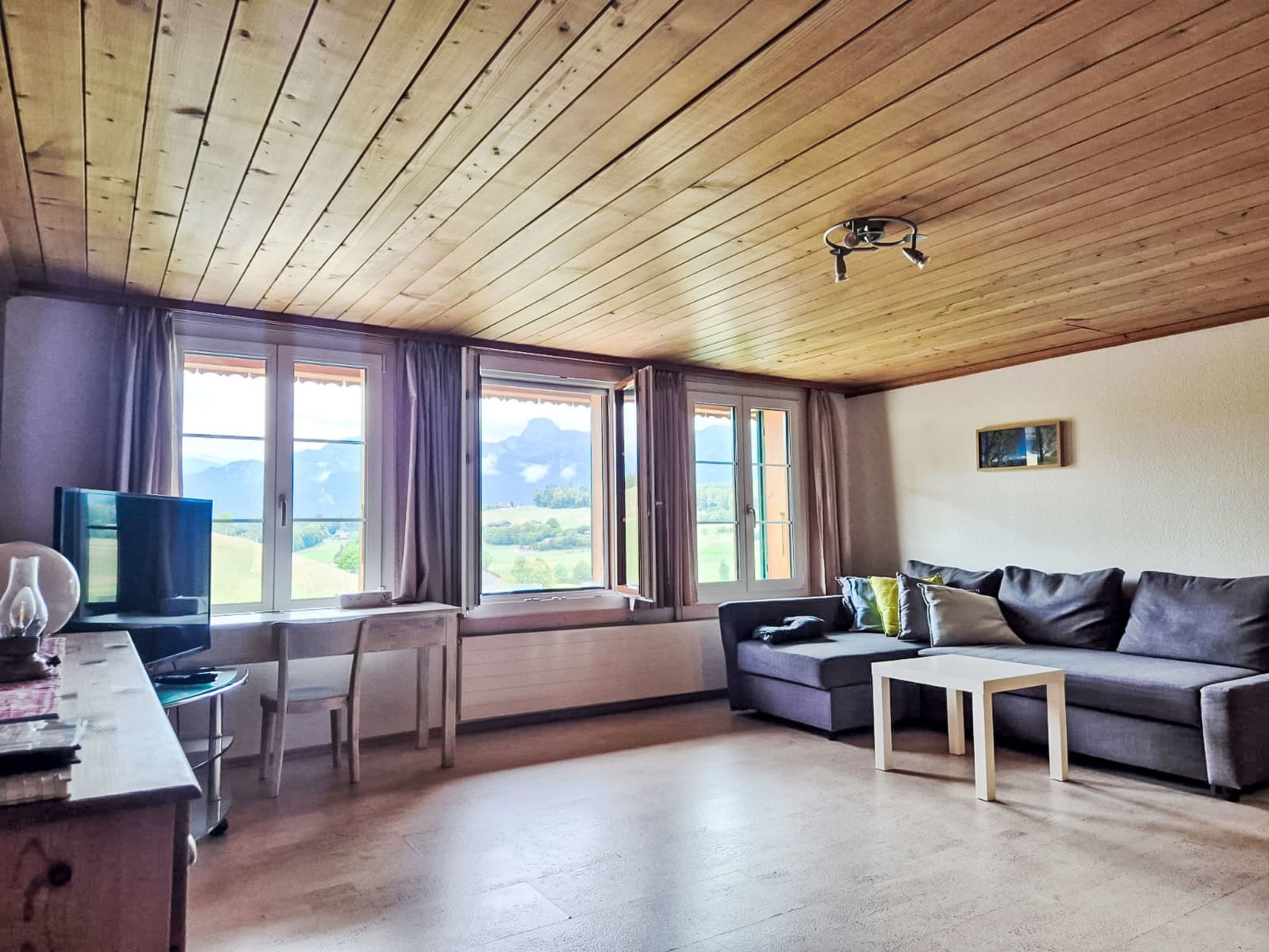 A room with a wooden ceiling, a sofa, a TV, and a view of mountains through large windows.