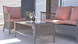 Een terras met rieten zitgedeelte, gewatteerde stoelen en een glazen tafel, met een fles en glazen.