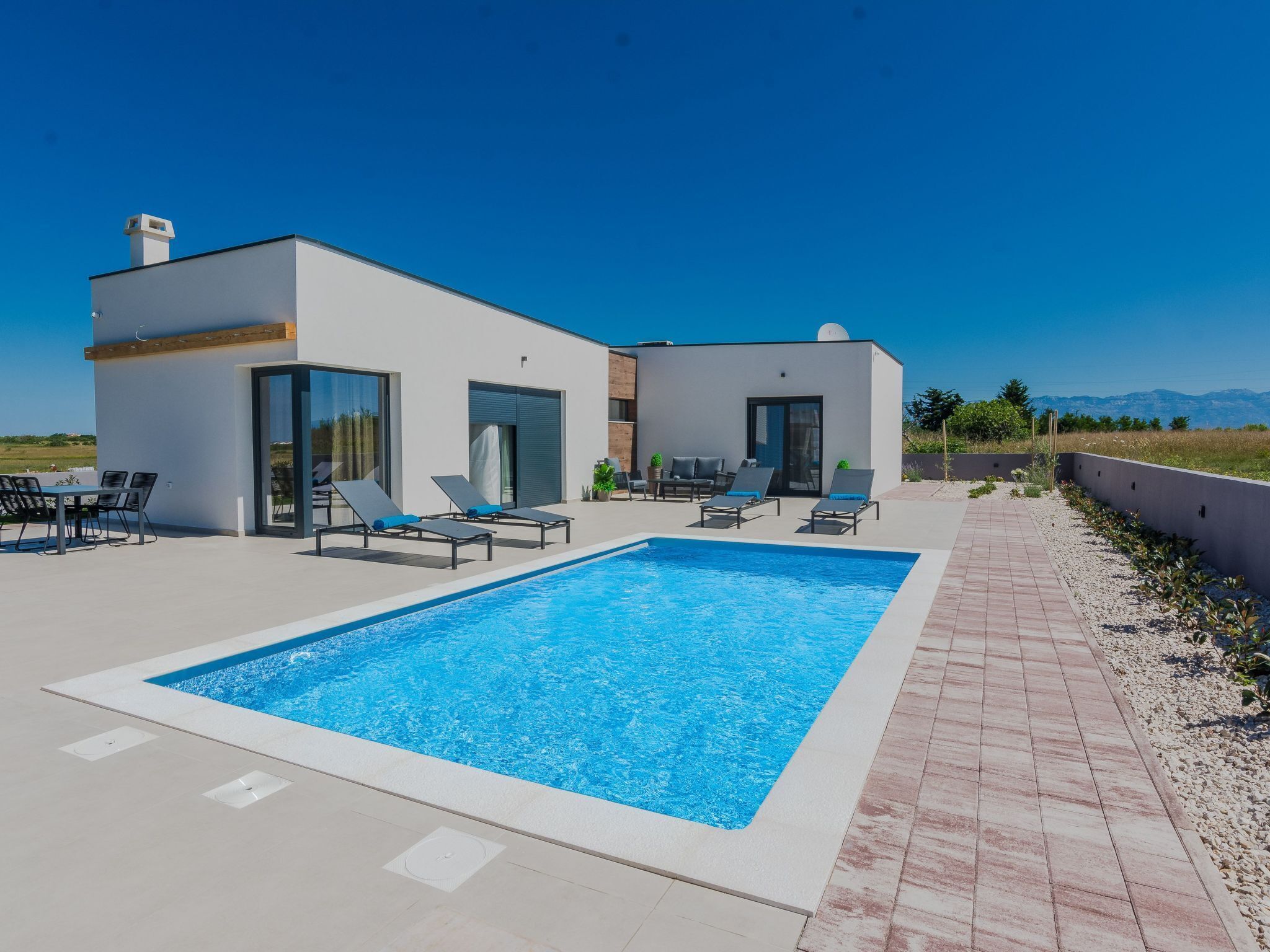 A modern outdoor pool area with a rectangular pool, lounge chairs, and a dining table.