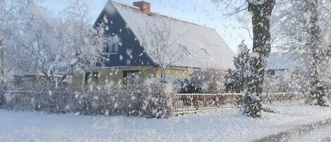 Une maison avec un toit en pente et une cheminée, entourée d’arbres et d’arbustes couverts de neige.