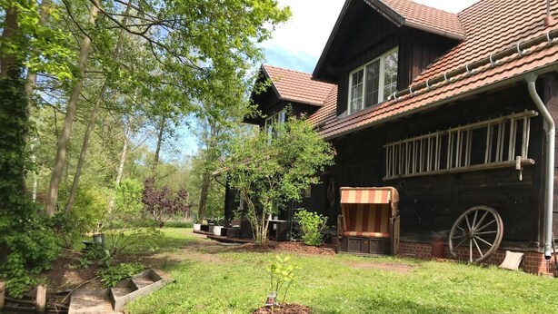 Una casa rustica in legno con facciata in mattoni, un molo in legno e un piccolo giardino con una carriola di legno e una panchina.