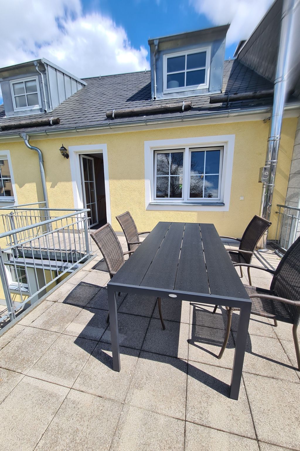 A patio with a table and chairs, a yellow building with a window, and a metal railing.