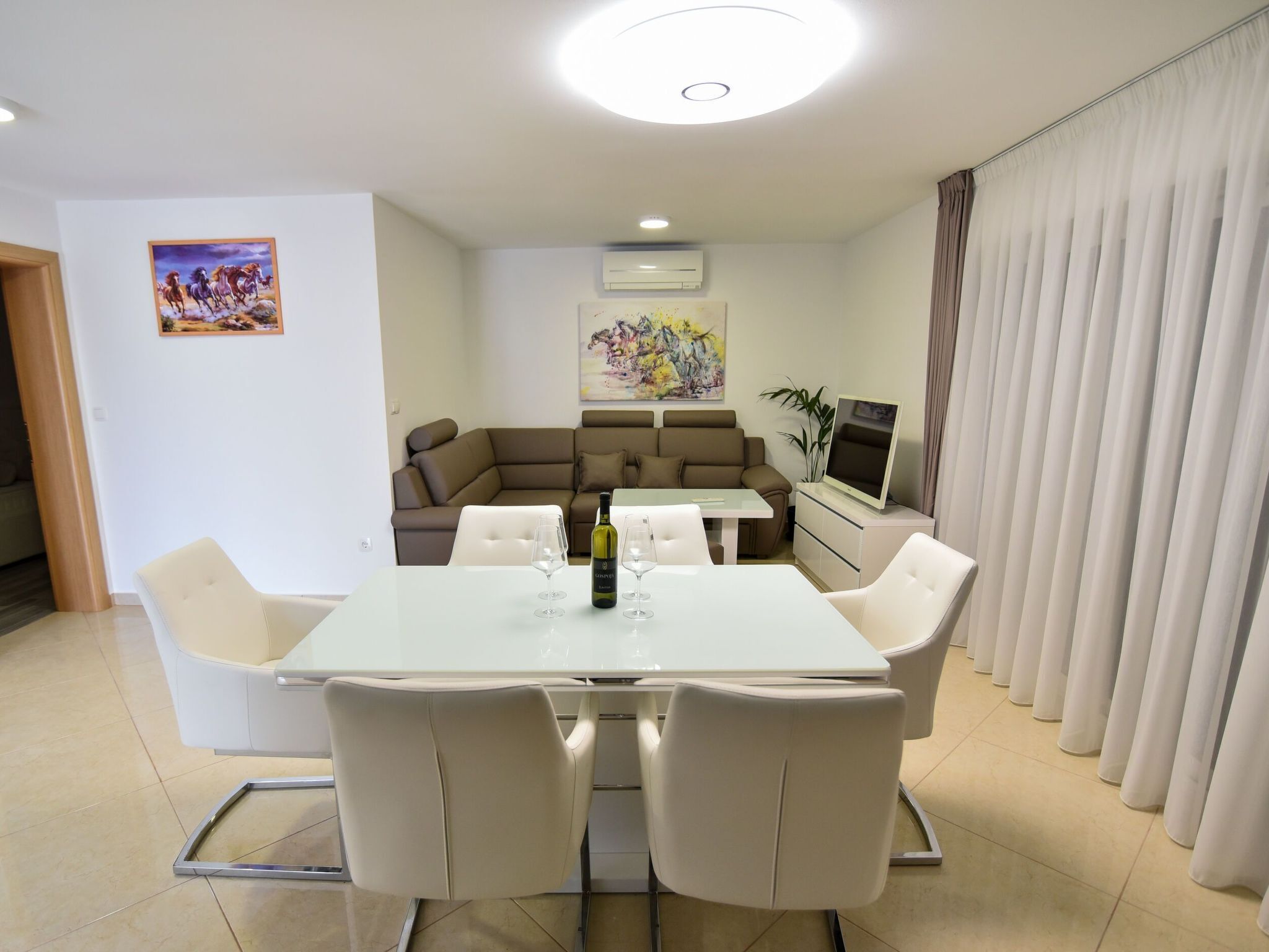 Una sala moderna con una mesa de comedor blanca, sillas color beige, un sofá y un televisor.