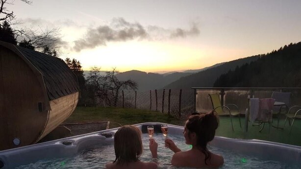 Dos personas en un jacuzzi con vistas a una puesta de sol y montañas.