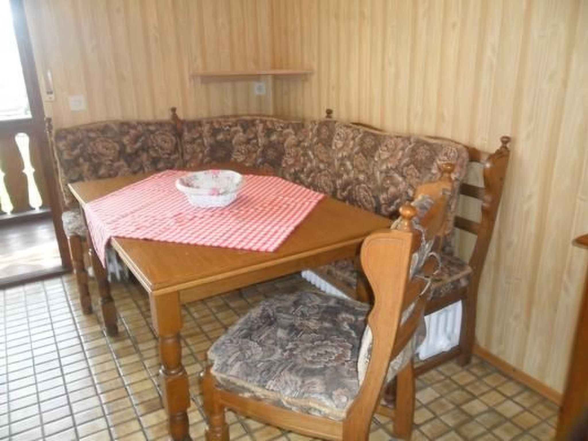 Ein Holzes Esstisch mit kariertem Tischläufer und einer Schüssel. Eine gemusterte Couch befindet sich dahinter.