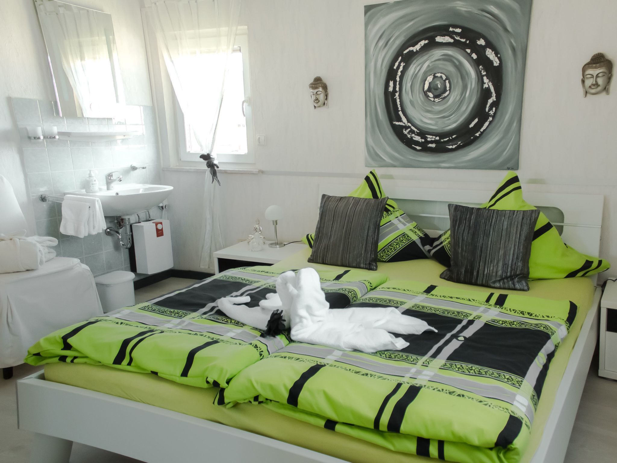 A modern bedroom with a bed, pillows, a nightstand, a lamp, a window, and a bathroom visible in the background.