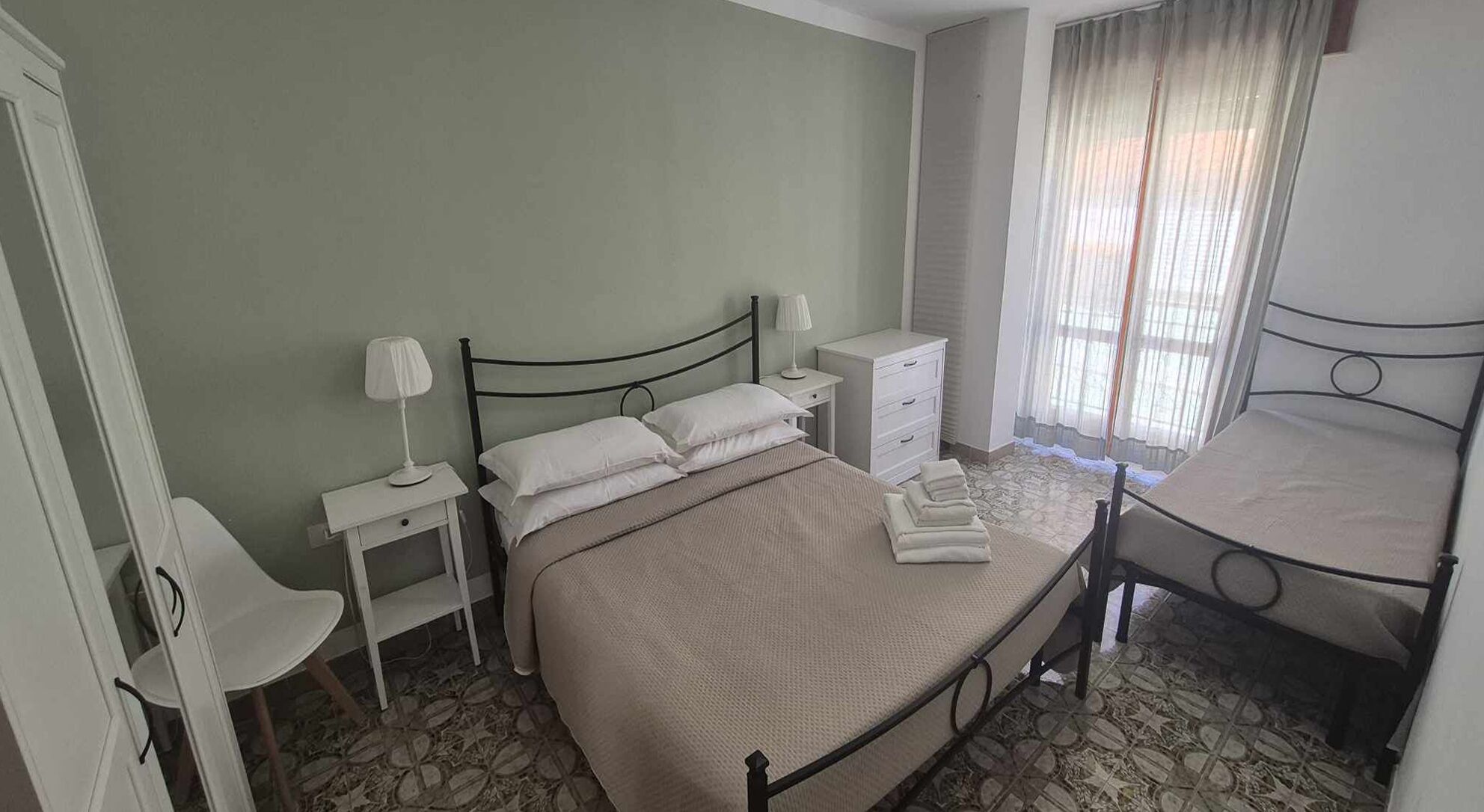 Une chambre à coucher avec un lit, des tables de chevet, une chaise, une commode et une fenêtre avec des rideaux.