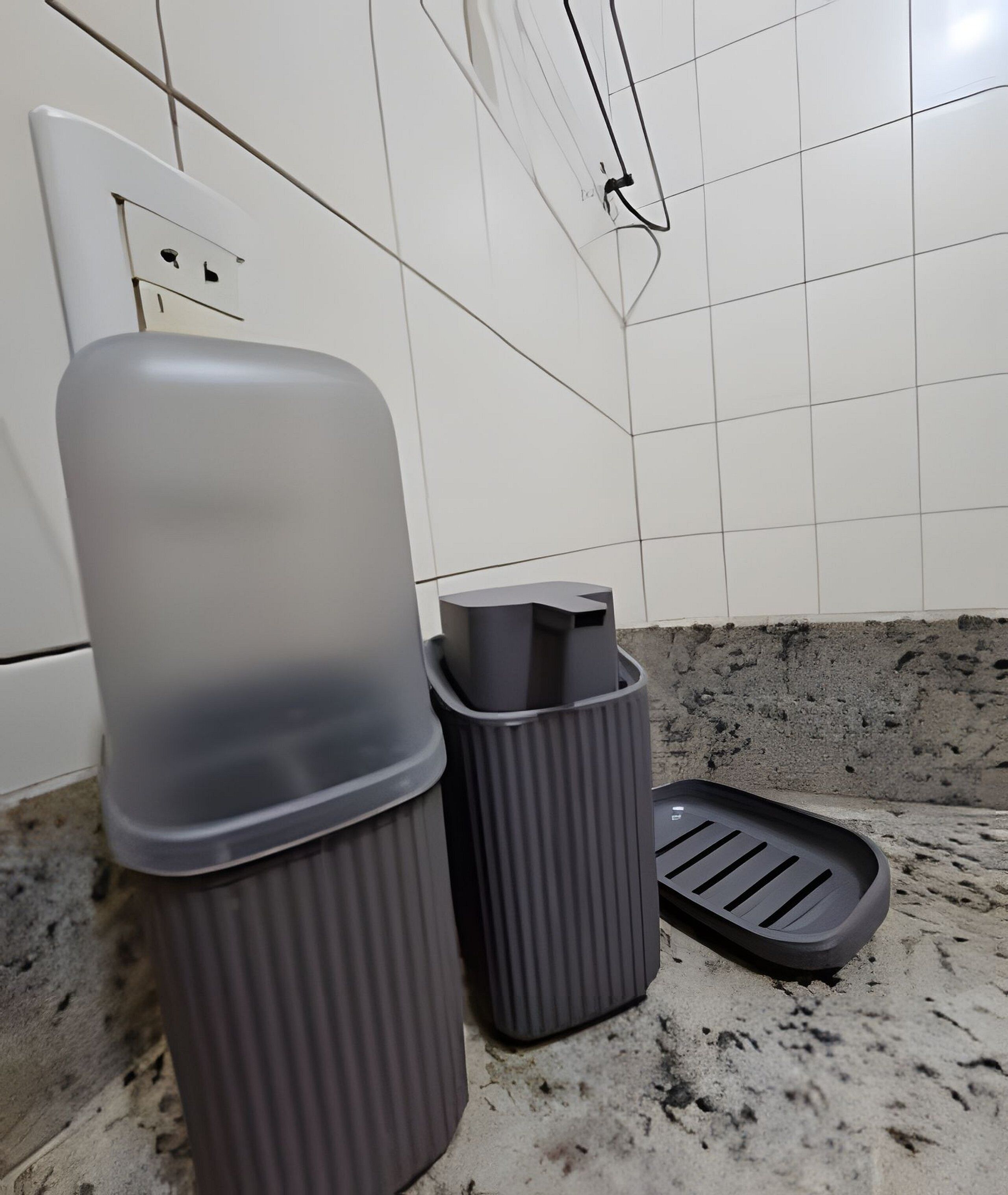 Une salle de bain avec un mur recouvert de carreaux blancs, une prise électrique blanche et deux poubelles grises.