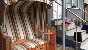 En træbænk med stribede hynder, et gelænder i metal og en potteplante på en balkon.