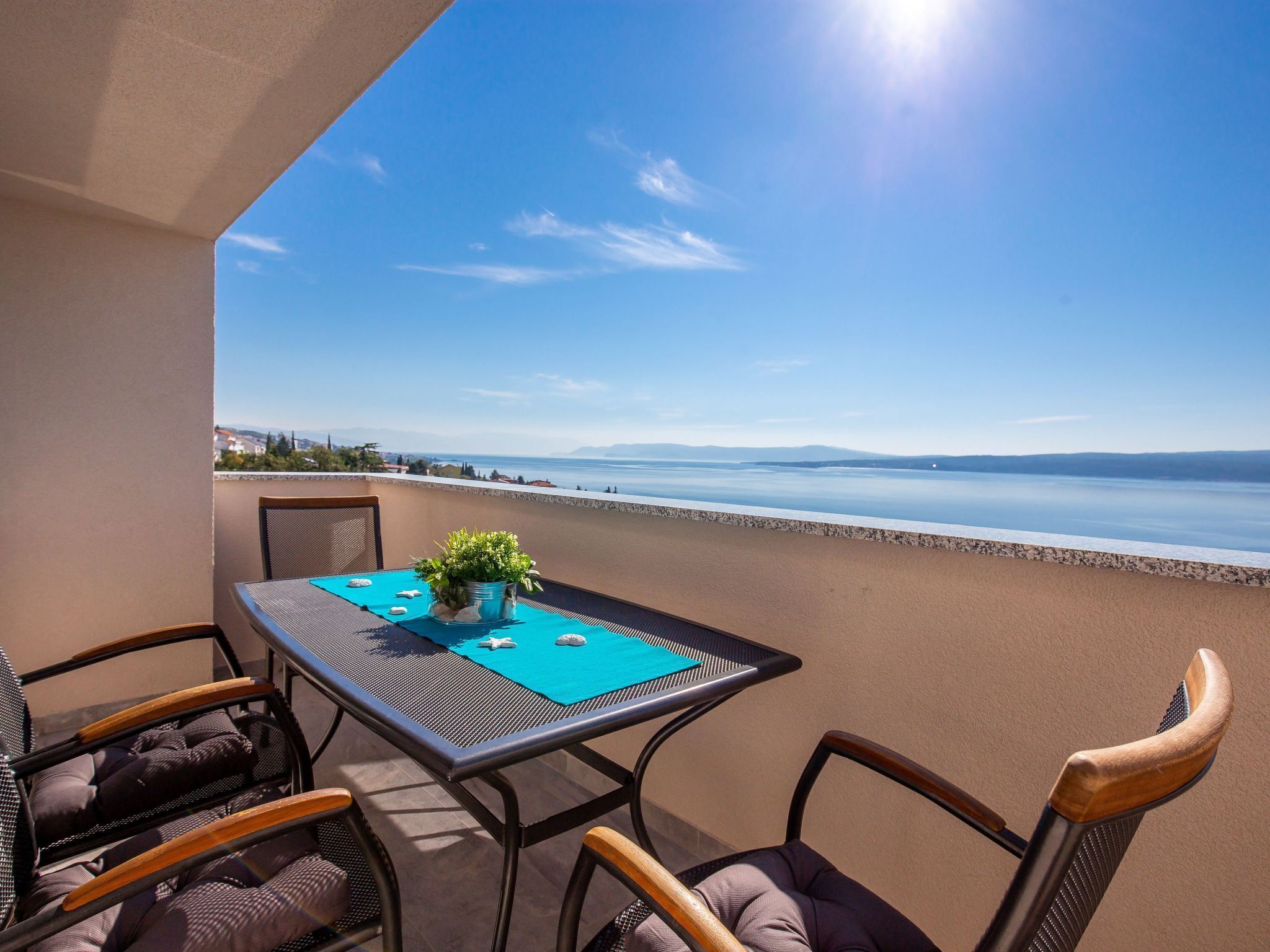 A balcony with a table and chairs, overlooking a calm sea and distant mountains.