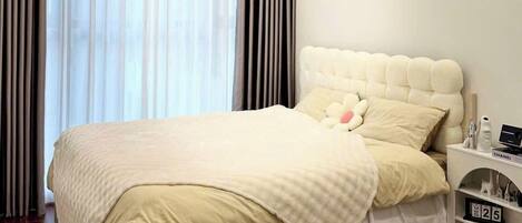 A modern bedroom with a large bed, a nightstand with a clock, and a window with sheer curtains.