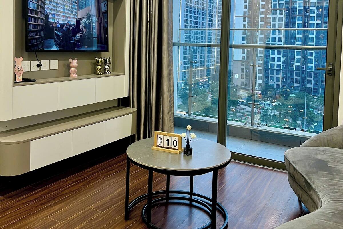 Ein modernes Wohnzimmer mit einem Flachbildfernseher, einem kleinen Tisch mit Datumsanzeige, einer Couch und einem großen Fenster mit Blick auf die Stadt.