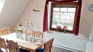 Een eetruimte met een houten tafel, blauwe gestoffeerde stoelen, een witte vaas met bloemen en een raam met rode gordijnen.