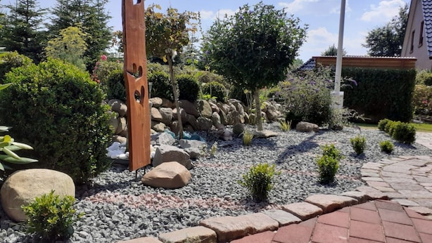 Een aangelegde tuin met een houten paal, rotsen en verschillende struiken.