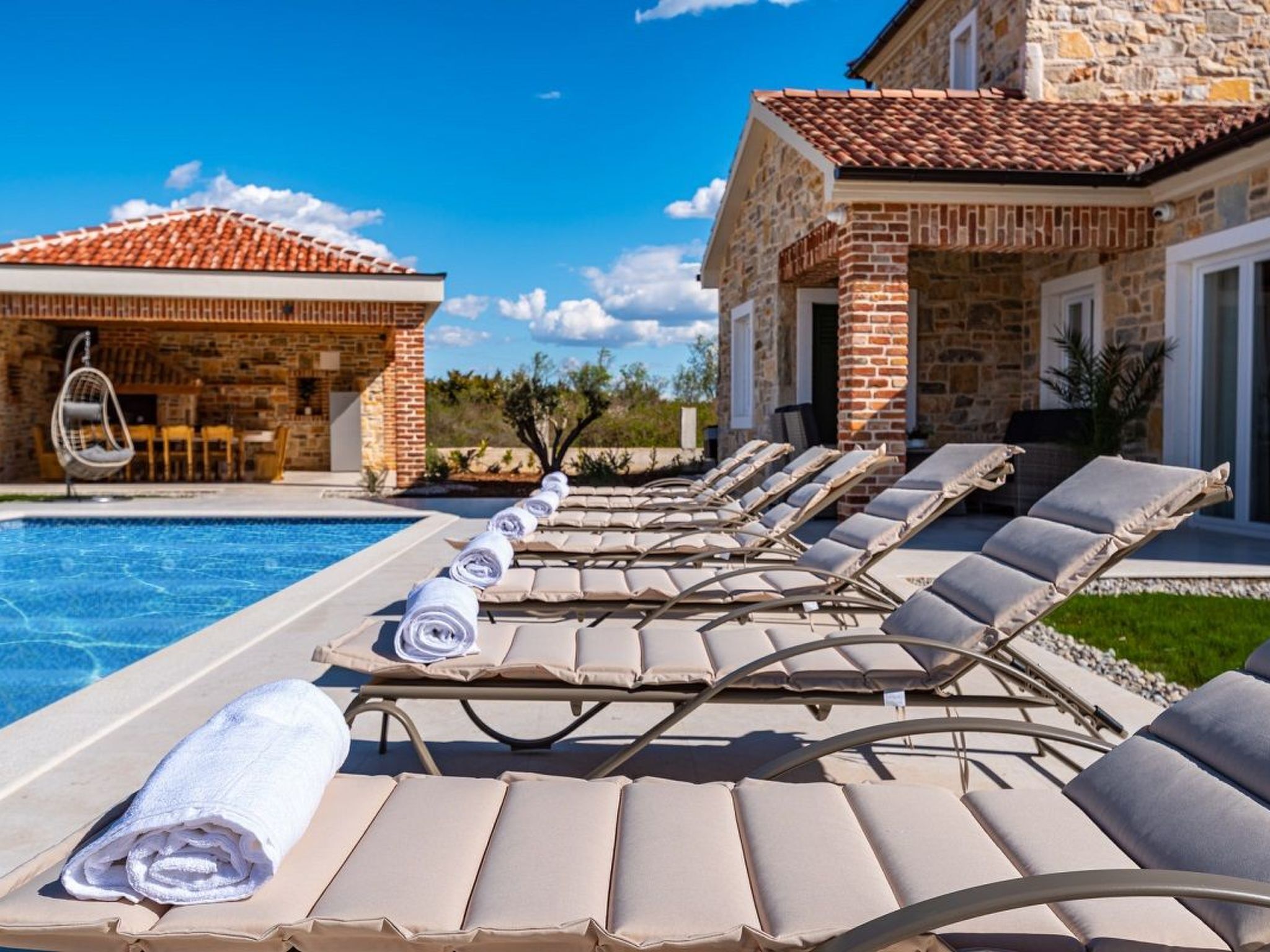 Un espace aménagé au bord de la piscine, avec des chaises longues et un bâtiment en pierre surmonté d’un toit en tuiles.