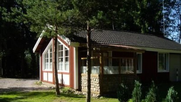 Ein kleines, eingeschossiges Haus mit Steinunterbau und Ziegeldach, umgeben von einem gepflegten Rasen und einigen jungen Bäumen.