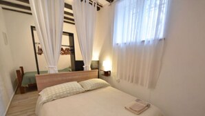Une chambre à coucher avec un lit, des rideaux blancs, une table de chevet avec une lampe et une fenêtre ornée de rideaux légers.