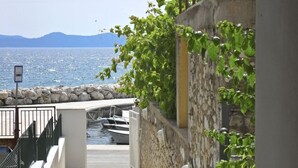 Un sentier de pierres menant à un quai où sont amarrés des bateaux, un mur de pierres recouvert de verdure et une vue dégagée sur la mer et les montagnes en arrière-plan.