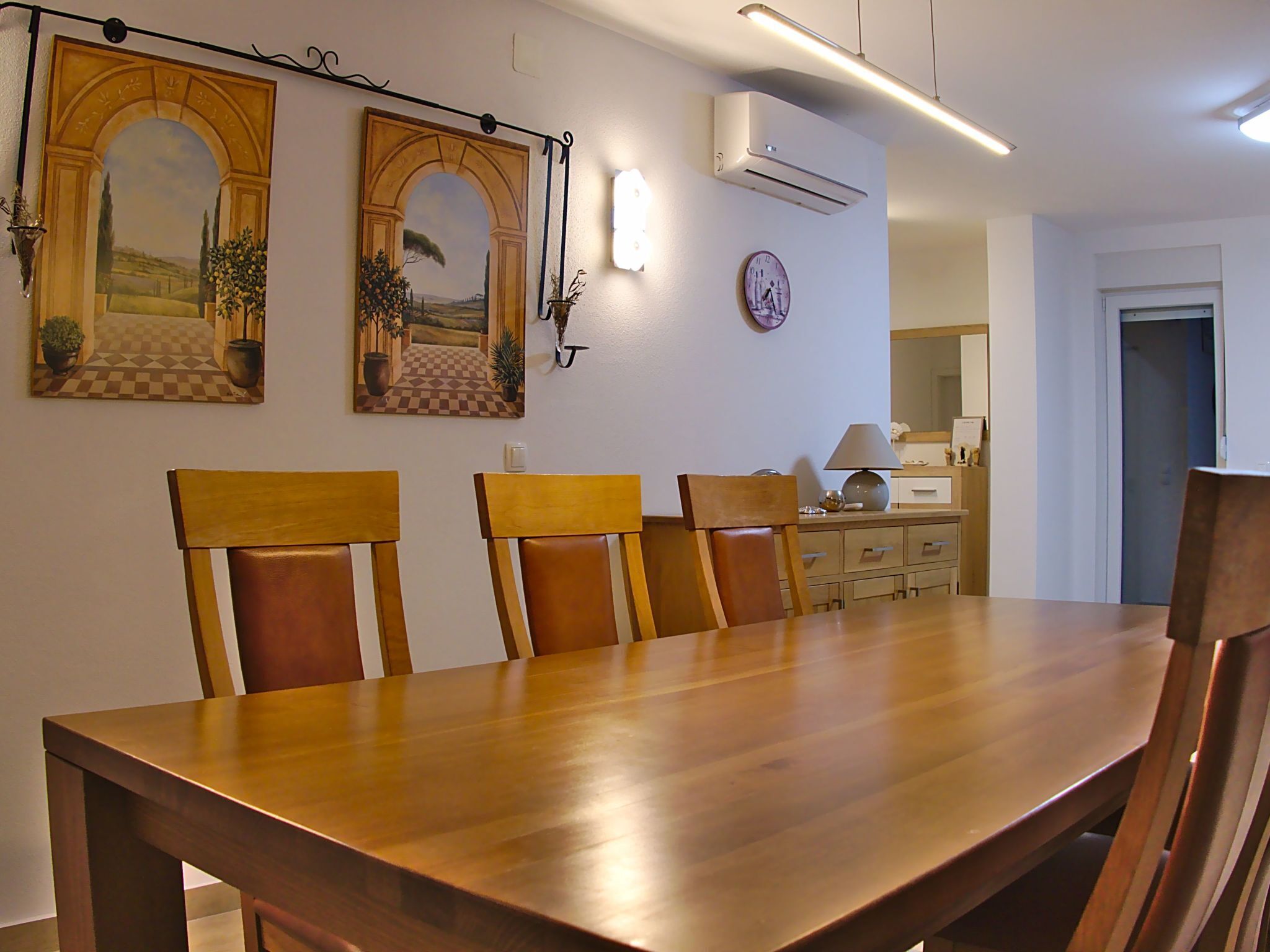Un coin repas avec une table et des chaises en bois, un climatiseur fixé au mur et deux tableaux encadrés.
