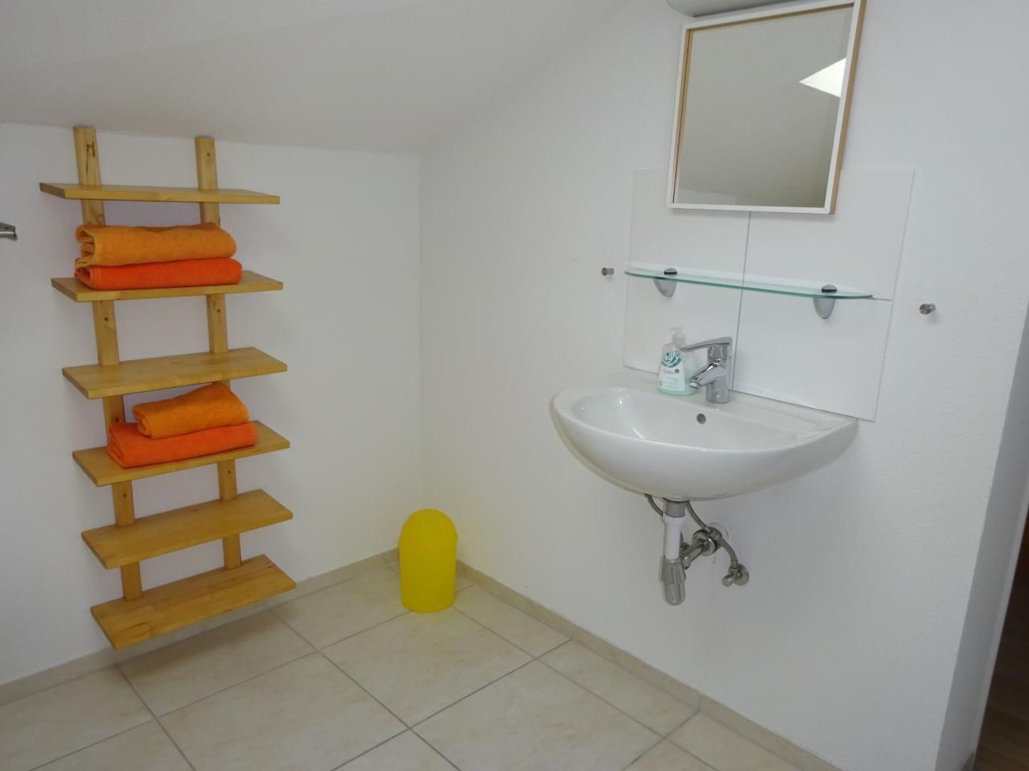 A bathroom with a wooden towel rack holding orange towels, a white sink with a glass shelf above it, and a mirror on the wall.