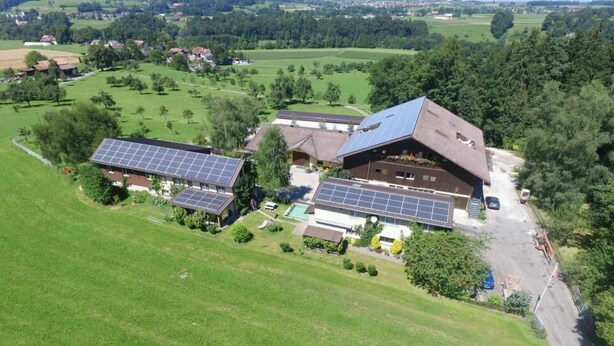 Ein großes Gebäude mit mehreren Solaranlagen auf dem Dach, umgeben von grünen Feldern und Bäumen.