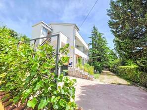 Ein zweistöckiges Gebäude mit Balkon, umgeben von Grünflächen und einer gepflasterten Auffahrt.