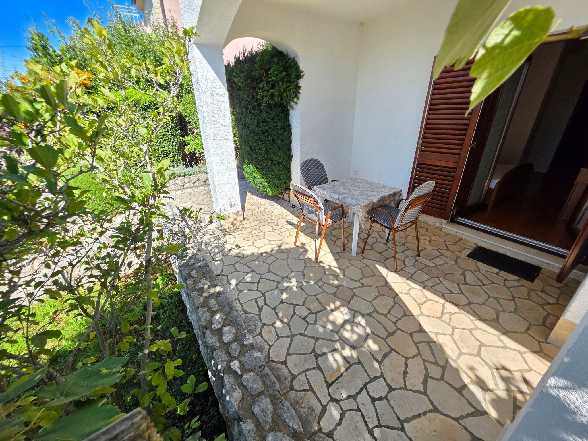 Un patio de piedra con una mesa y sillas, rodeado de follaje y una pared blanca.