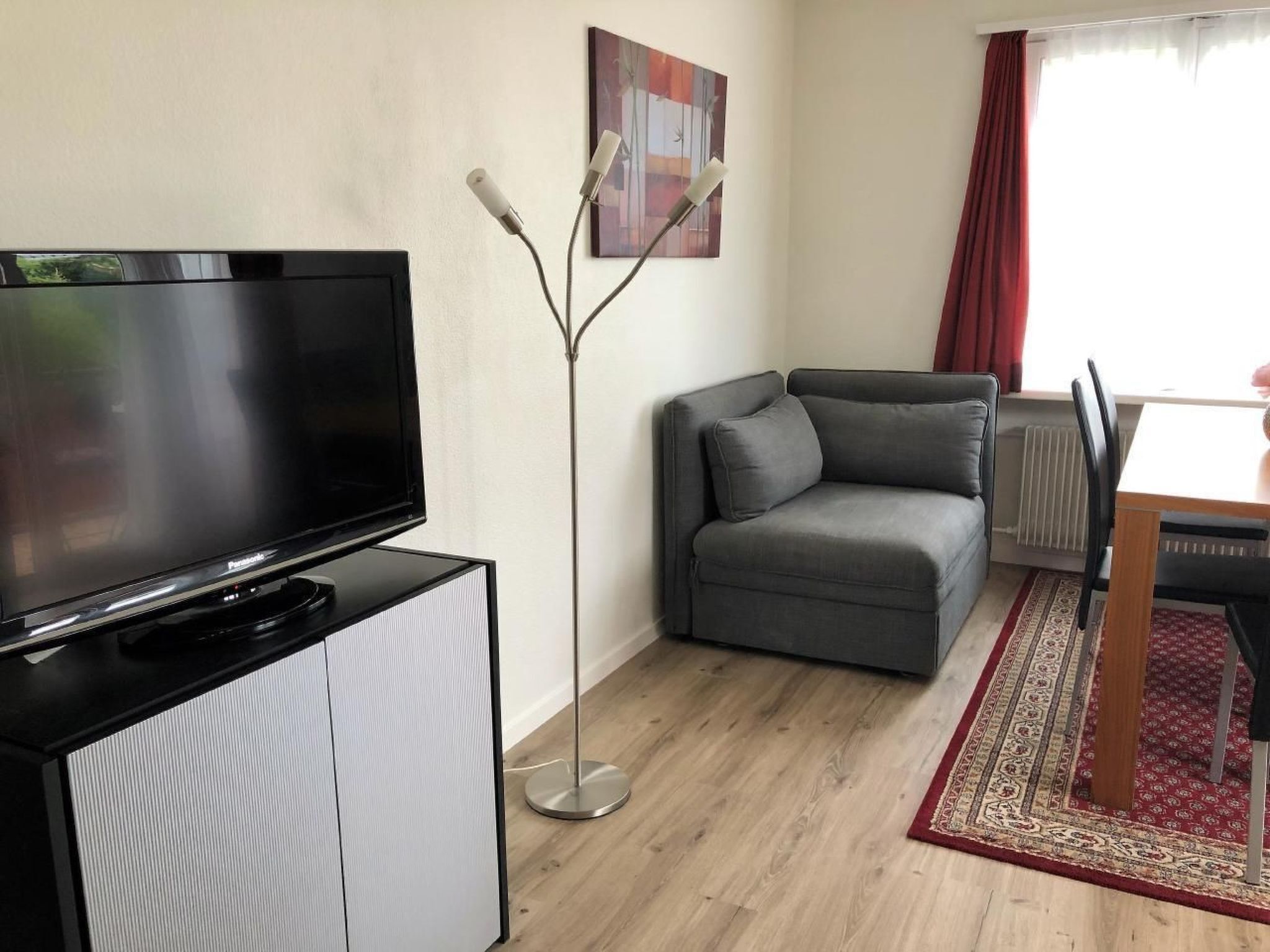 Een moderne woonkamer met een flatscreen televisie op een kast, een grijze bank, een vloerlamp en een eethoek met een tafel en stoelen.