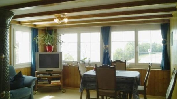 Ein Zimmer mit Esstisch, Stühlen, einem Fernseher auf einem Schrank, einer Couch und einem Fenster mit Vorhängen.