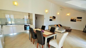 Een moderne keuken met witte kastjes en roestvrijstalen apparatuur, een eetgedeelte met een houten tafel en stoelen, en een woonkamer met een bank en een televisie.