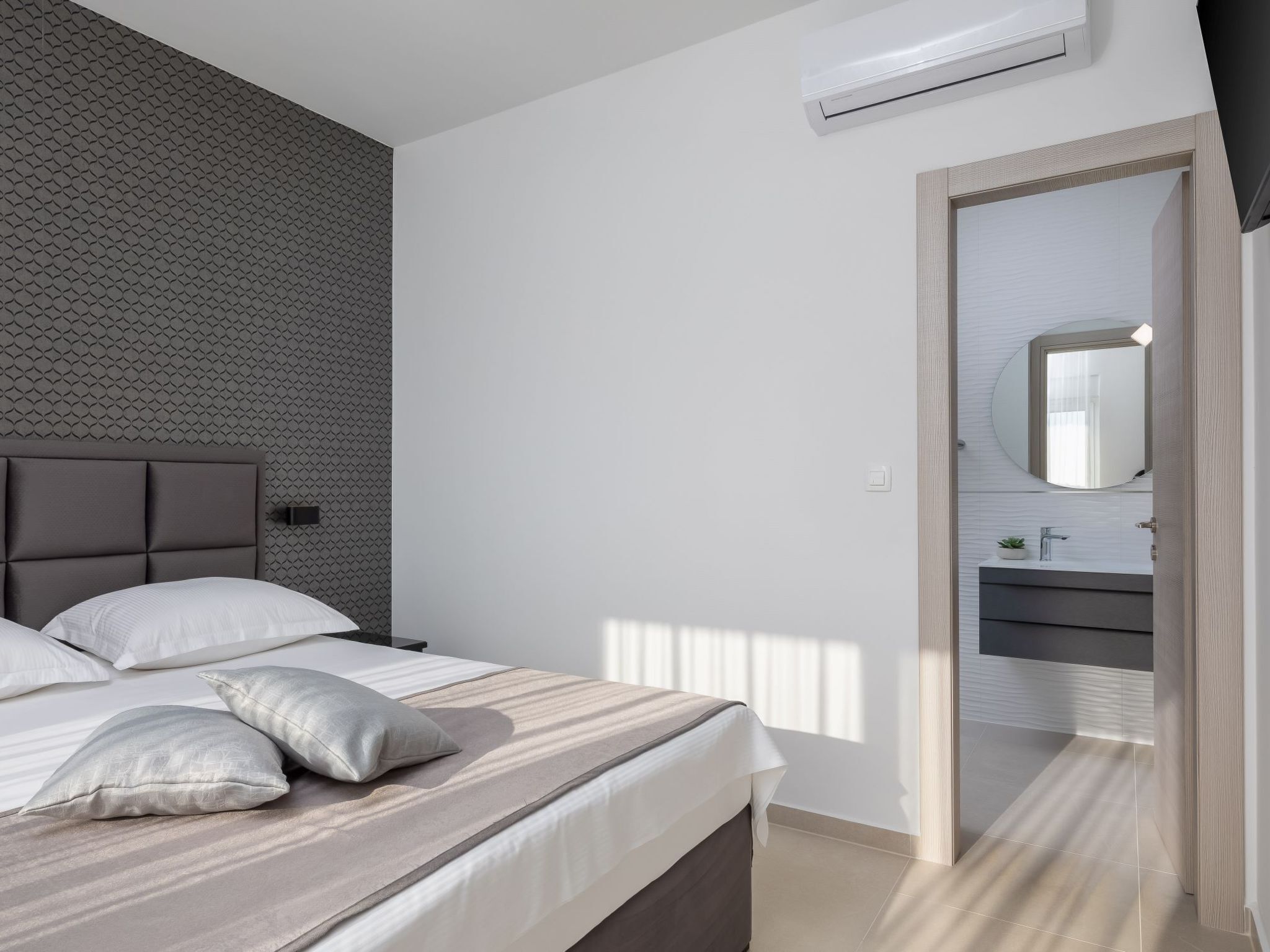 A modern bedroom with a bed, pillows, a headboard, an air conditioning unit, a bathroom with a sink and mirror, and a patterned wall.