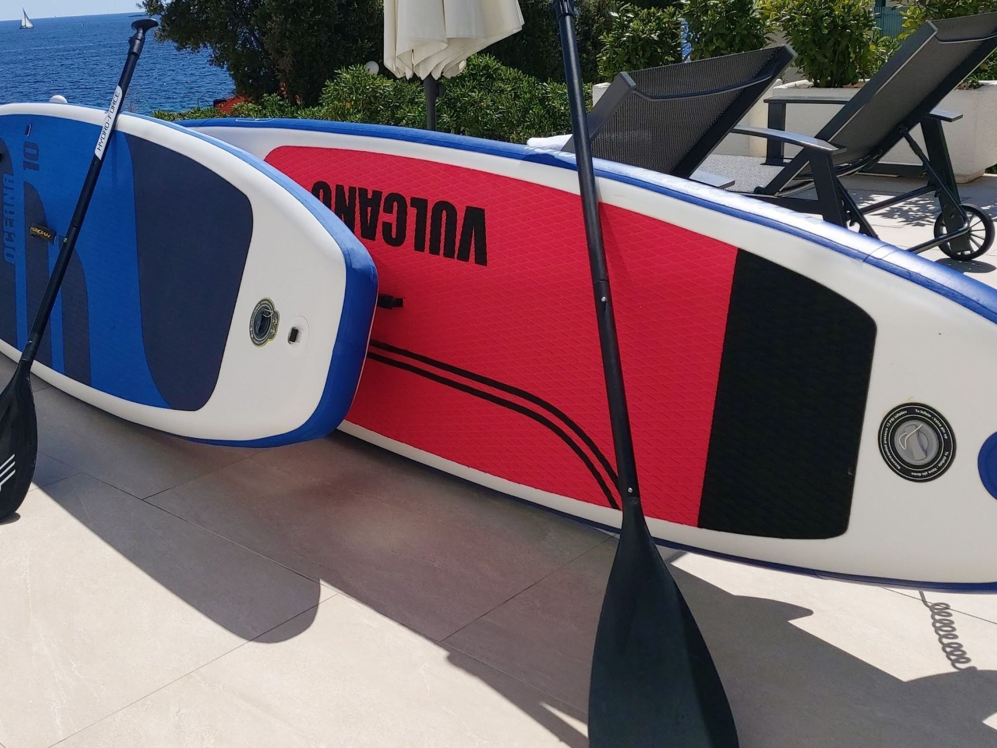 A paddleboard with the word "VOLKAN" on it, placed on a tiled surface near a lounge area with chairs and a table.