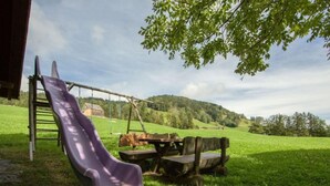Un parc infantil amb tobogan i cadira de balanceig a un camp de gespa, amb una casa al fons.