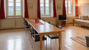 Een ruime kamer met een lange houten tafel, meerdere stoelen, een bed, een televisie en grote ramen met rode gordijnen.