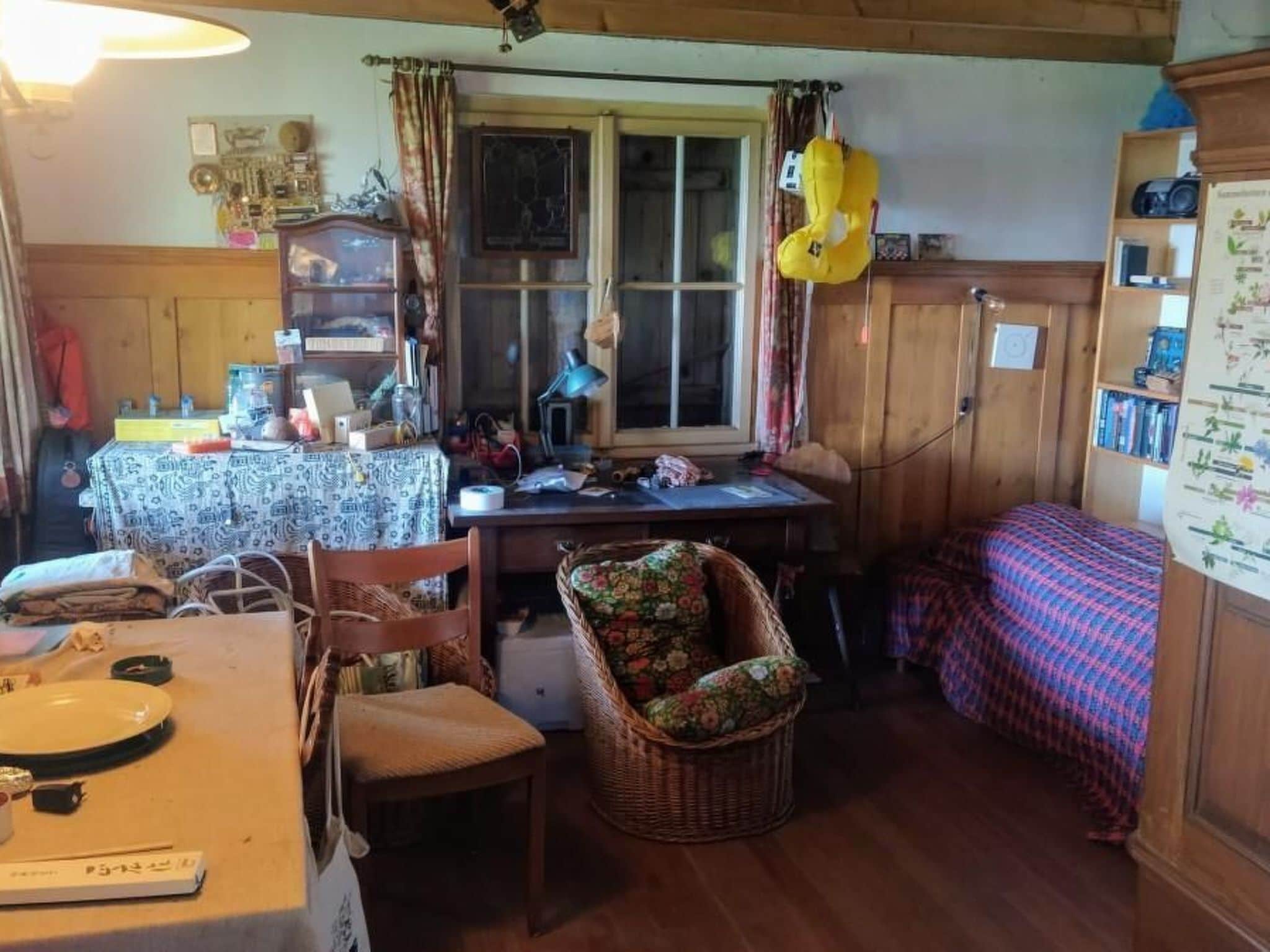 Une pièce chaleureuse avec une table à manger, une chaise, un panier en osier, un lit recouvert d’une couverture à carreaux et une fenêtre avec des rideaux.