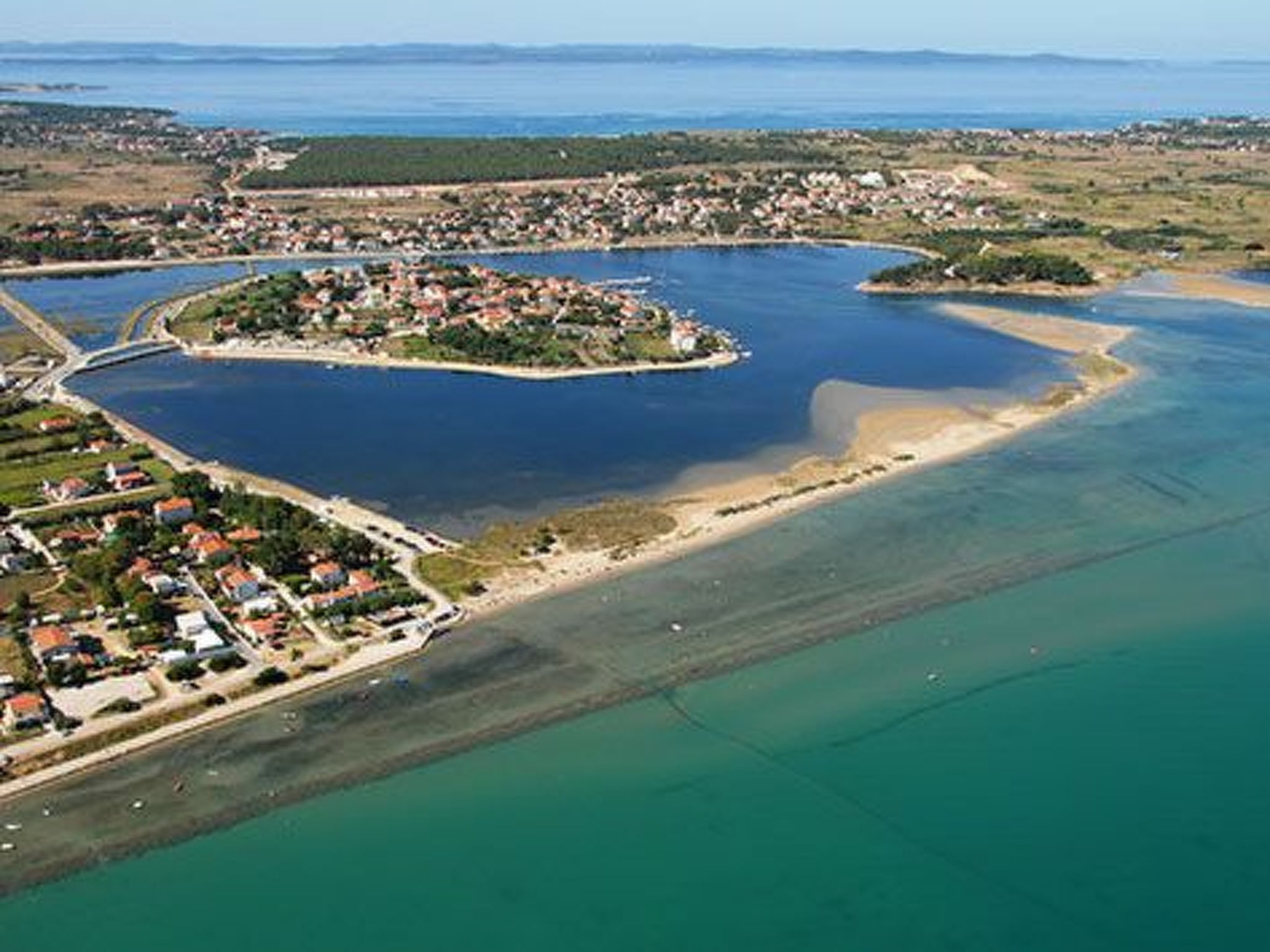 An aerial view of a coastal area with a small island featuring a cluster of buildings, surrounded by clear blue waters and a sandy beach.